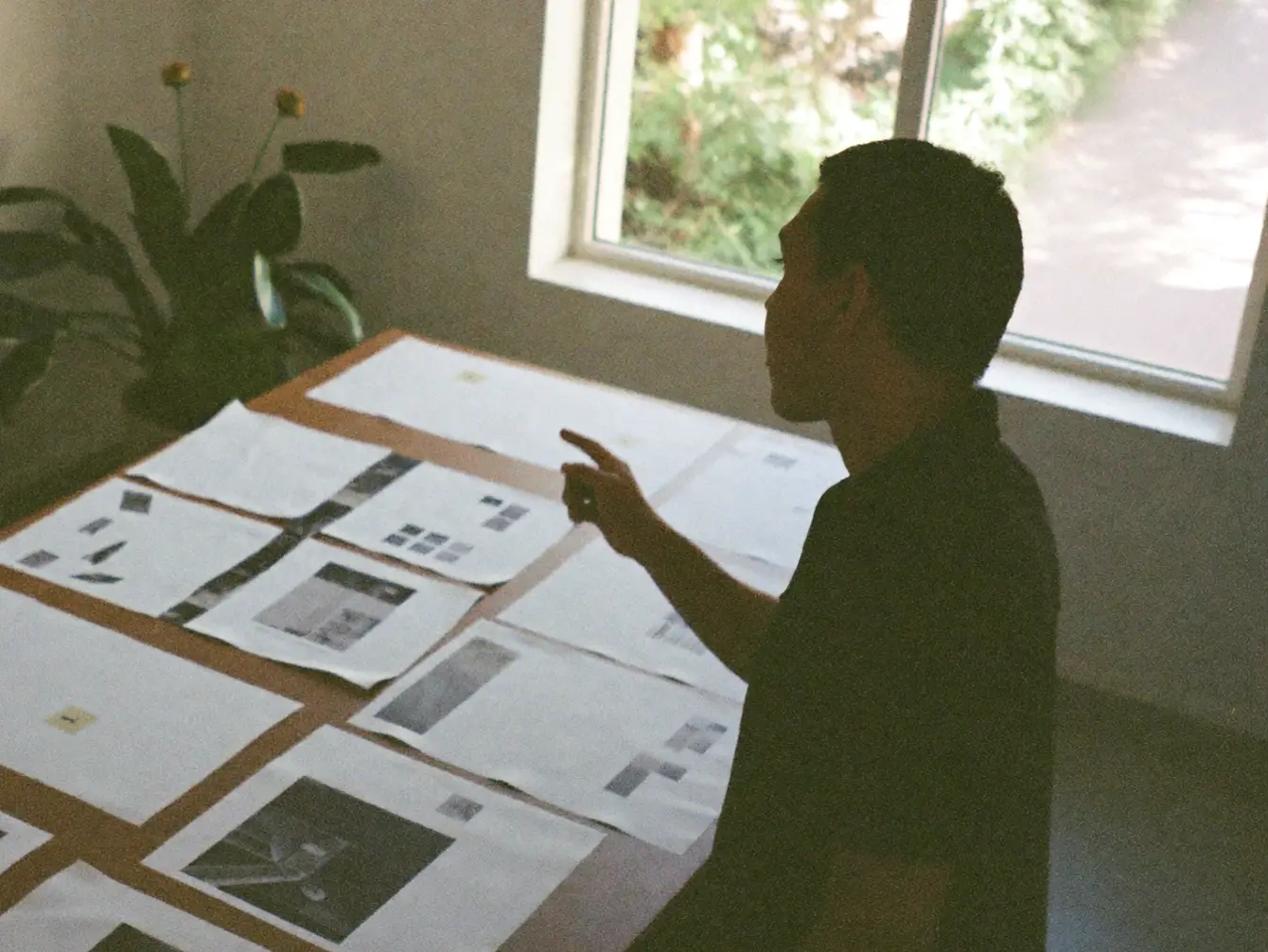 Someone leaning over a long table covered in printed page layouts, beside a tall window.