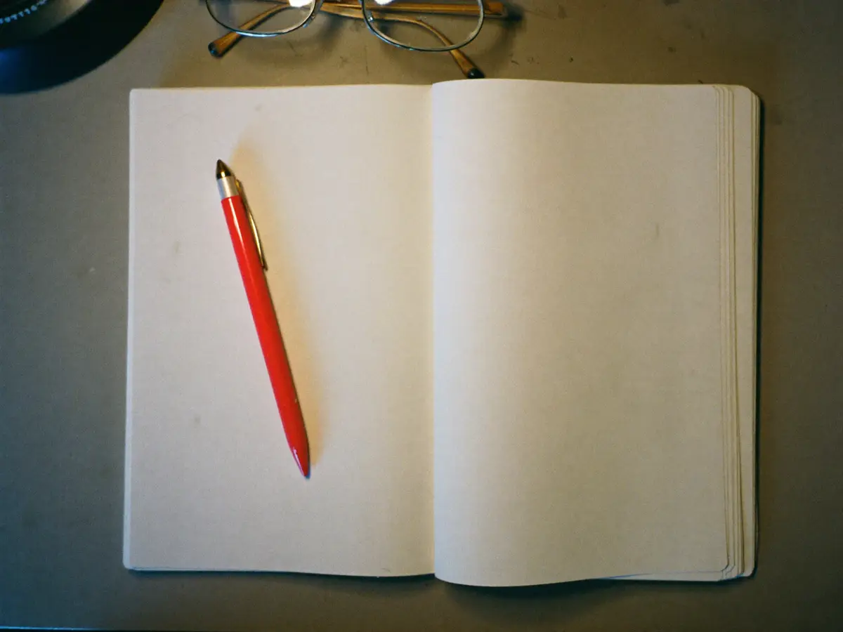 An open blank notebook with a red pen resting on it, under a desk lamp.