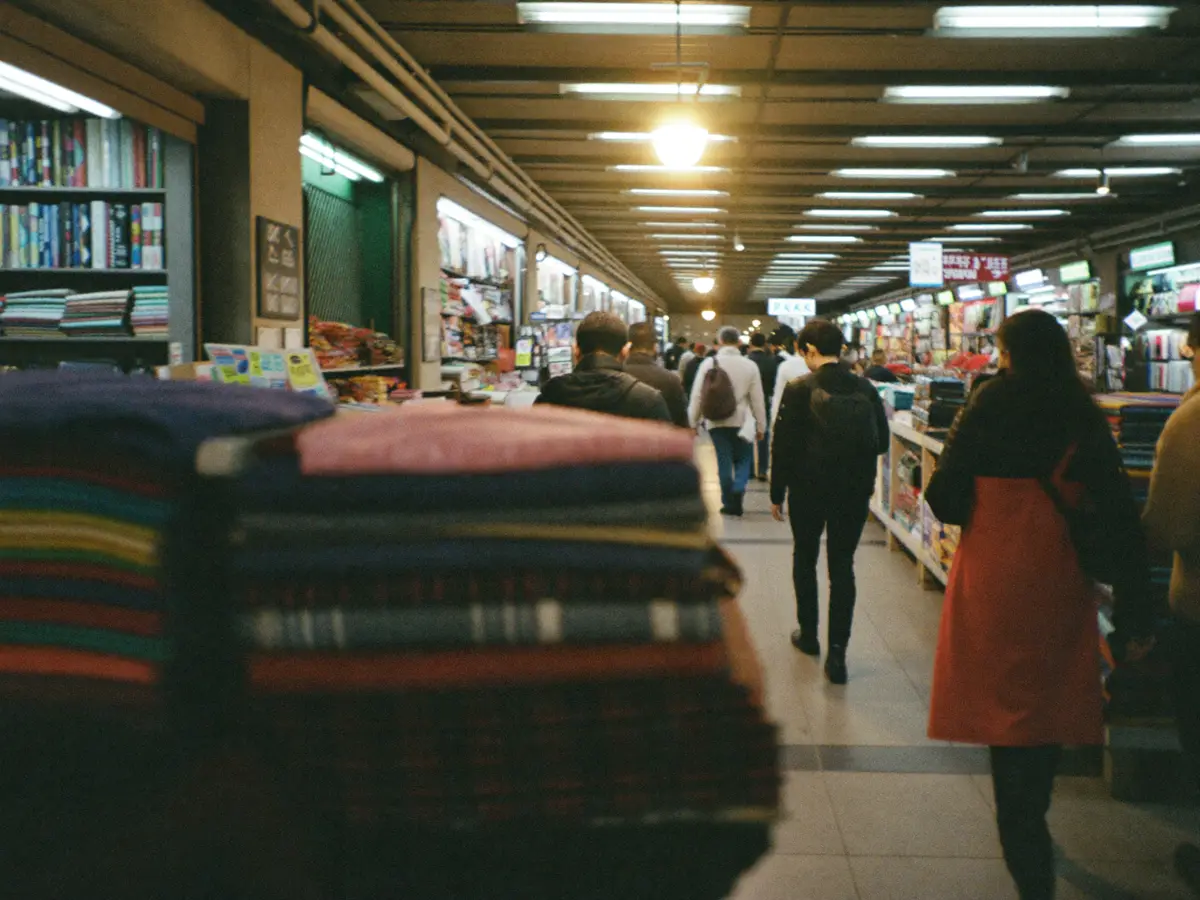 Shoppers moving through a busy covered market aisle past a stall of folded fabrics.