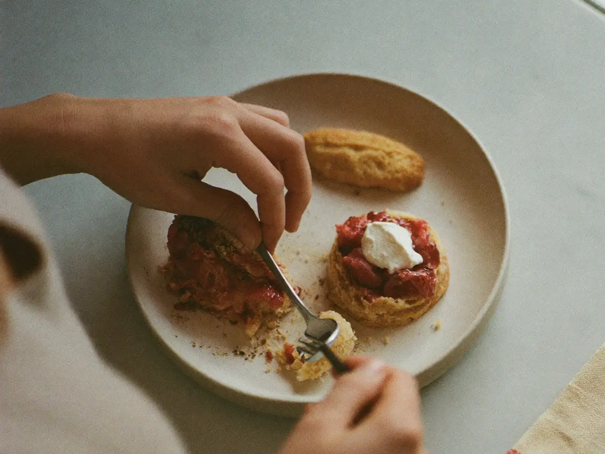 Hands finishing a dessert plate on a pale counter, seen from above.